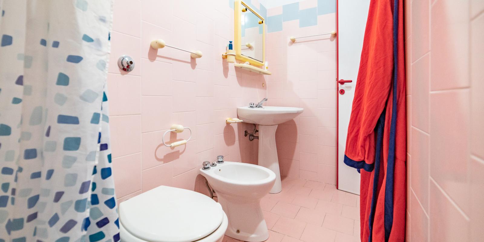 Bagno con piastrelle rosa, sanitari bianchi e tenda da doccia a motivi blu.