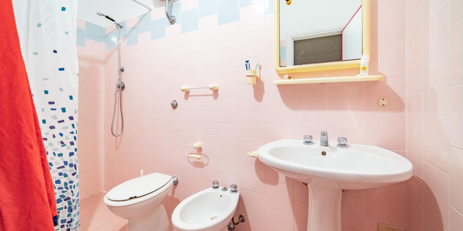 Bagno con piastrelle rosa, doccia, bidet, lavandino e specchio con mensola.