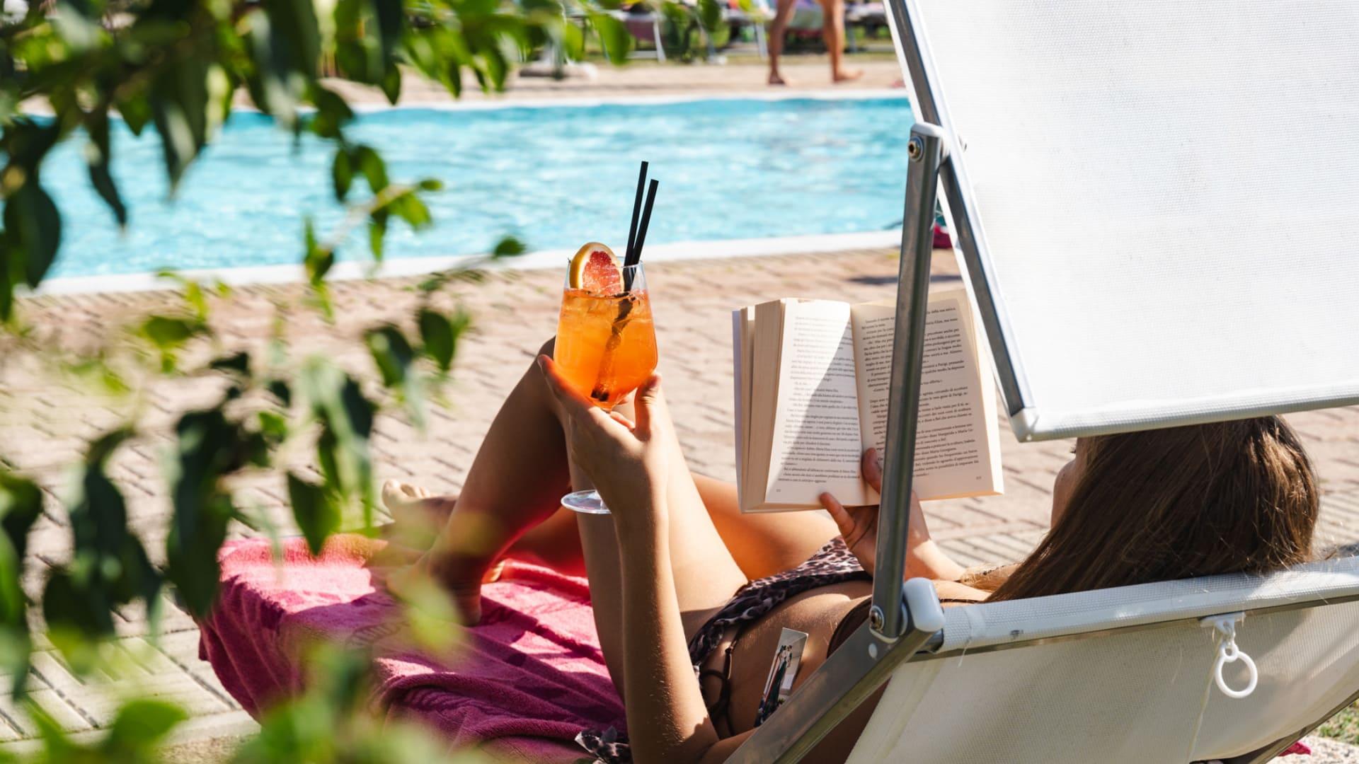 Donna su sdraio, cocktail in mano, legge un libro vicino alla piscina.