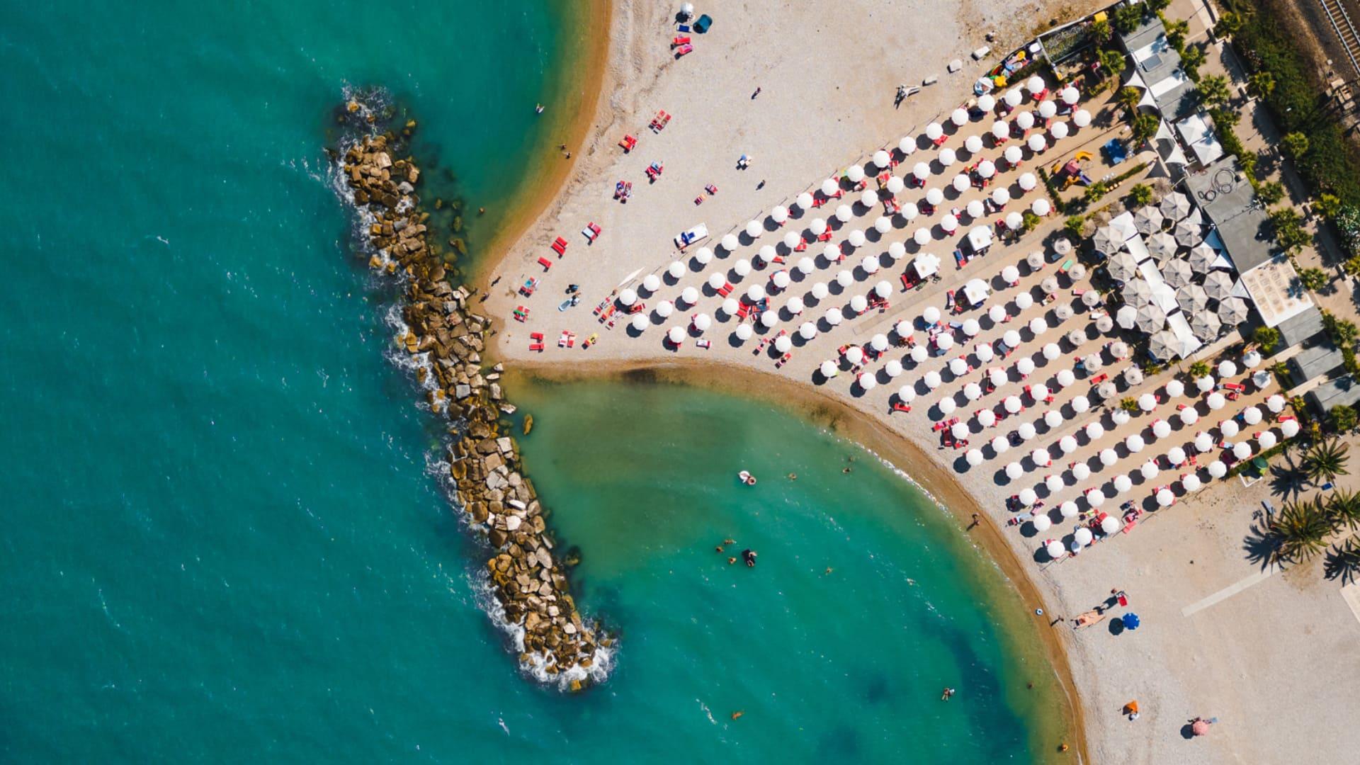 Spiaggia con ombrelloni bianchi e lettini rossi vista dall'alto, mare turchese e frangiflutti.