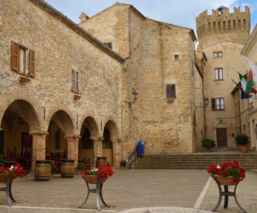 Piazza storica con archi, fiori rossi e una torre medievale in pietra.